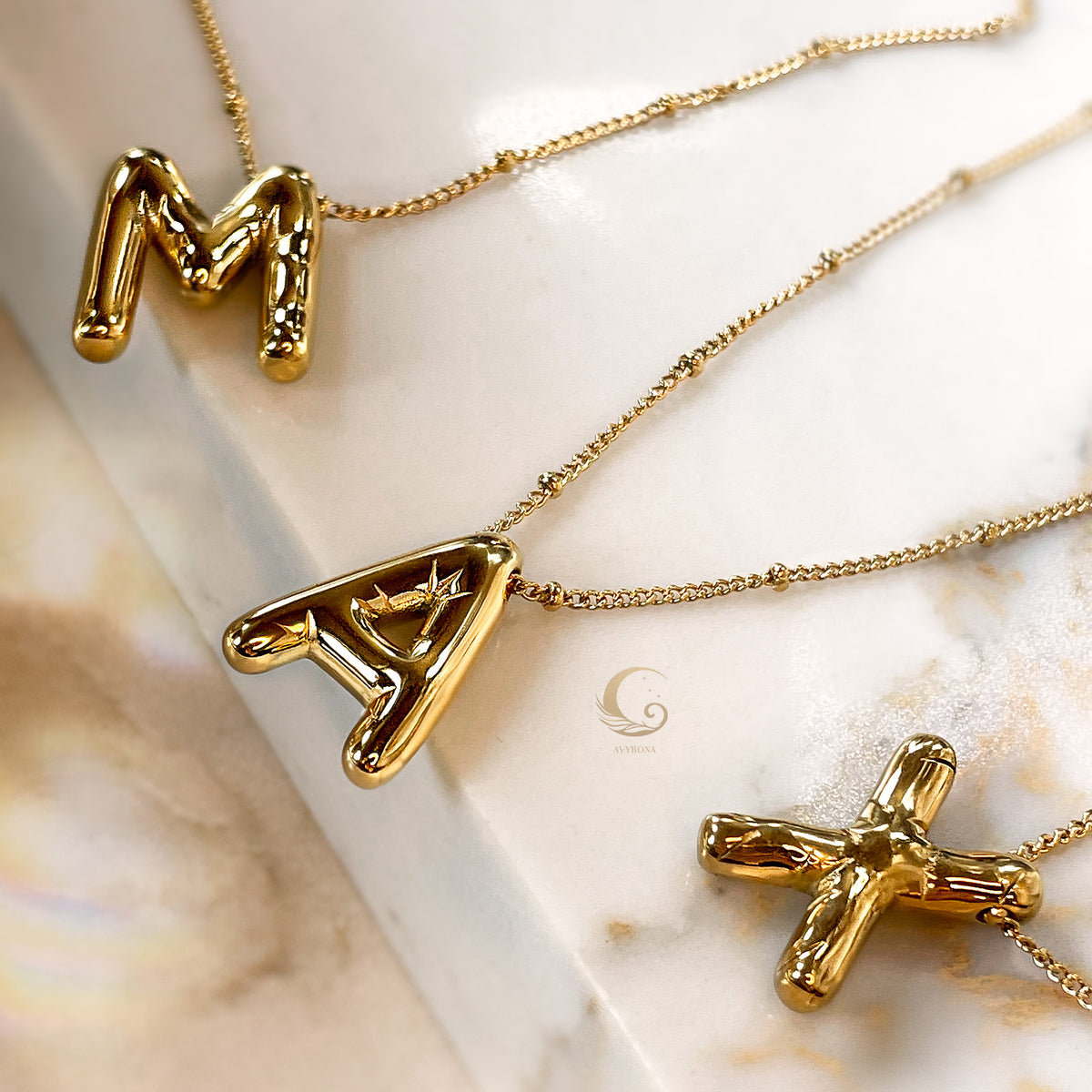 Gold balloon-style letter necklaces “M”, “A”, and “X” styled diagonally on a light marble surface.