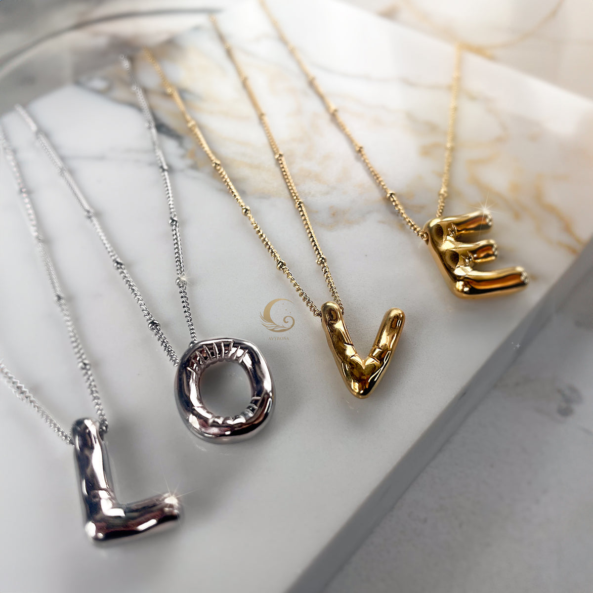 Four balloon-style letter necklaces spelling the word “LOVE” in mixed silver and gold finishes, laid on marble surface.