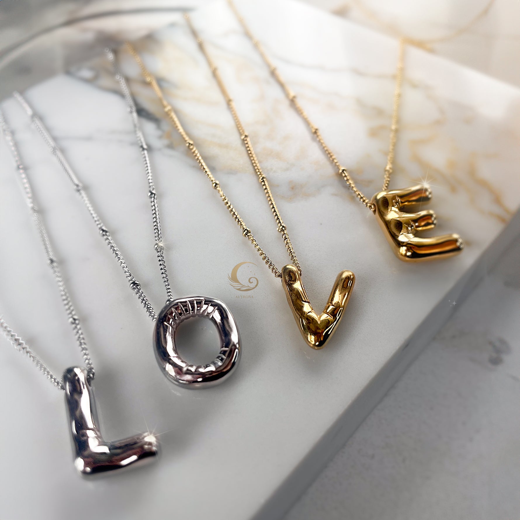 Four balloon-style letter necklaces spelling the word “LOVE” in mixed silver and gold finishes, laid on marble surface.