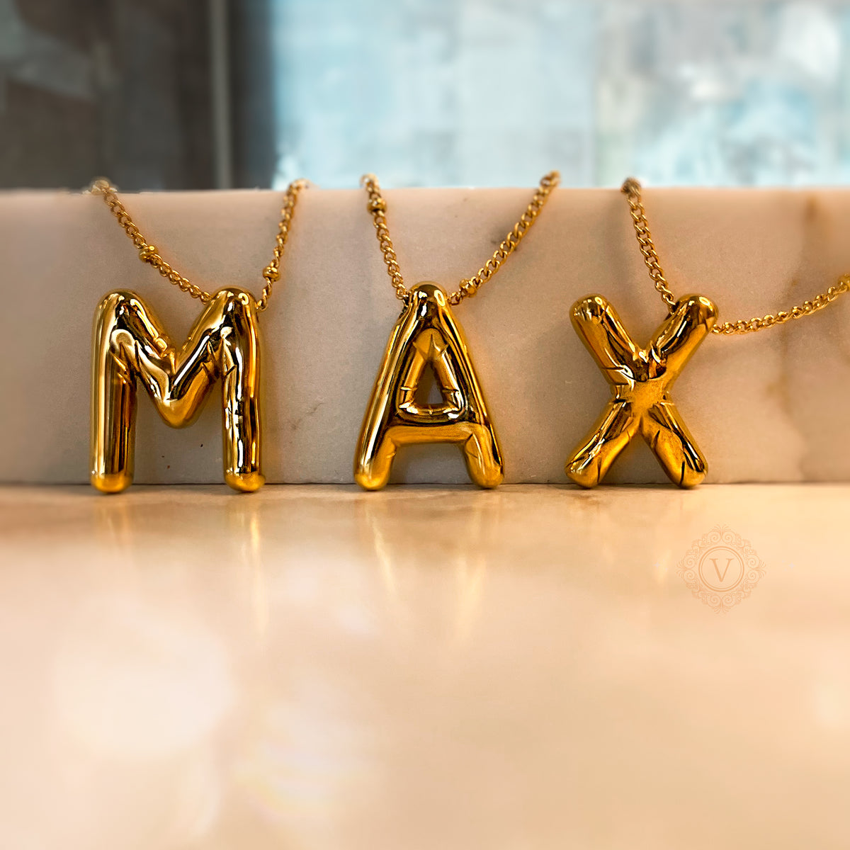 Three gold balloon letter necklaces spelling “MAX”, standing upright on soft beige surface with blurred background.