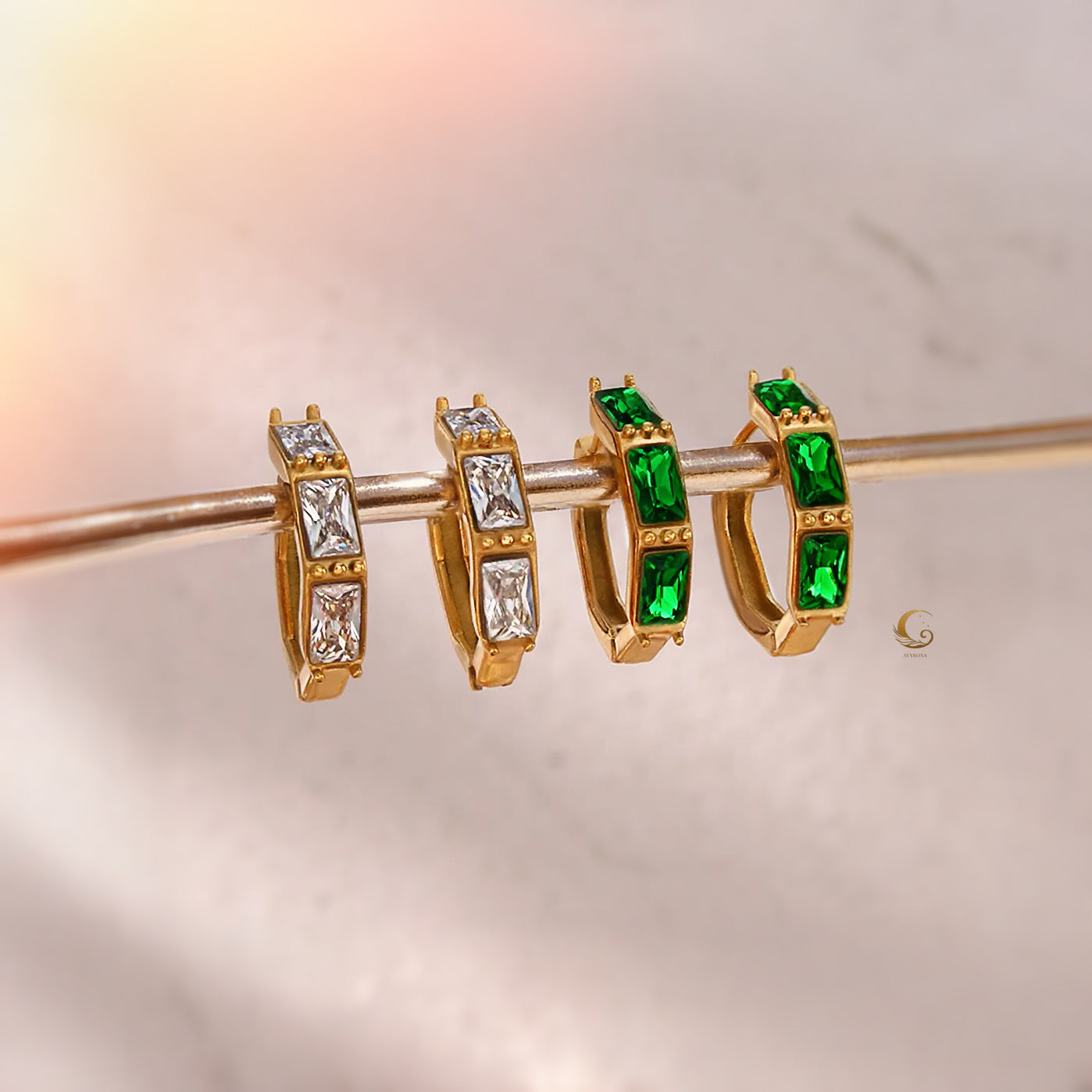 Four Camila hoop earrings displayed on a gold rod, showing two emerald green and two clear zircon stone variants