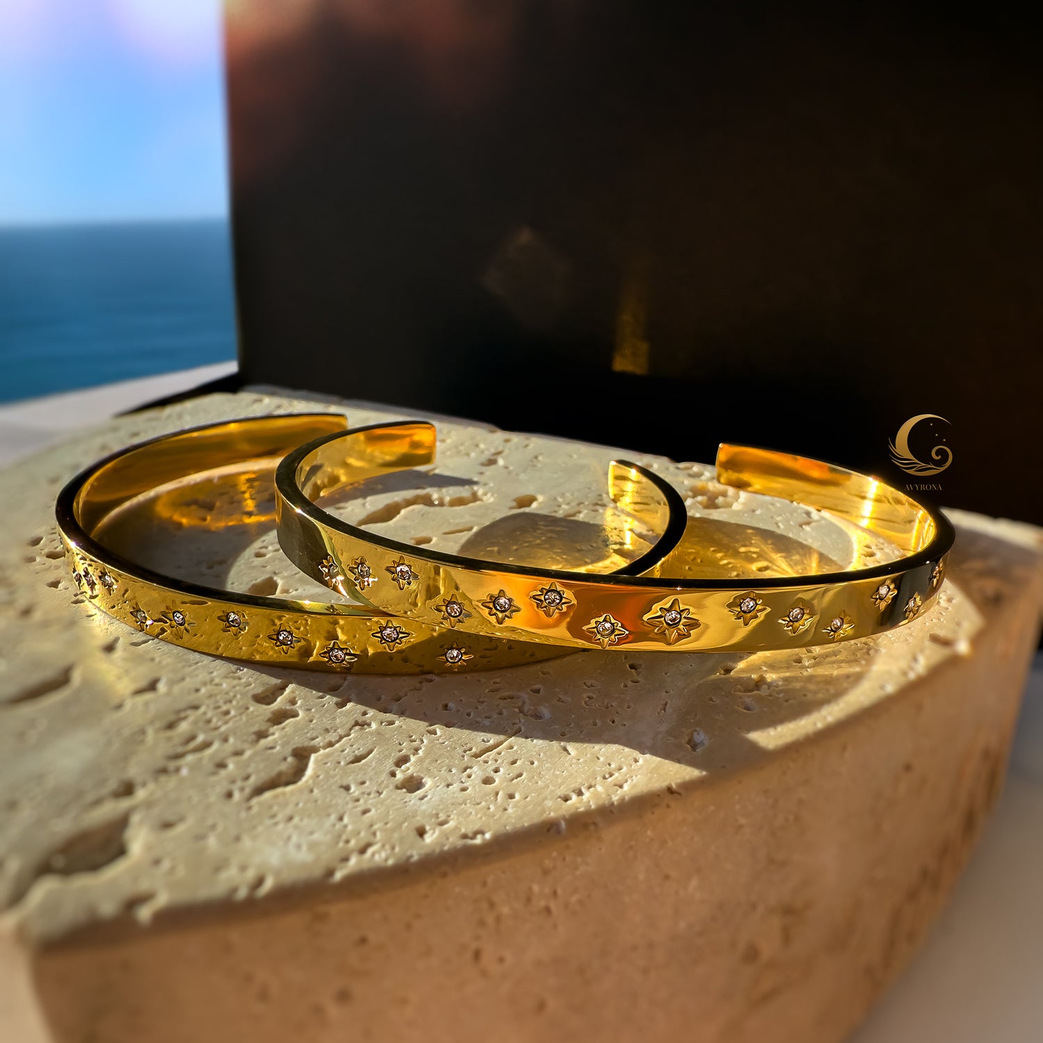 Two gold cuff bracelets with celestial star engravings and cubic zirconia, resting on a natural stone under warm light.