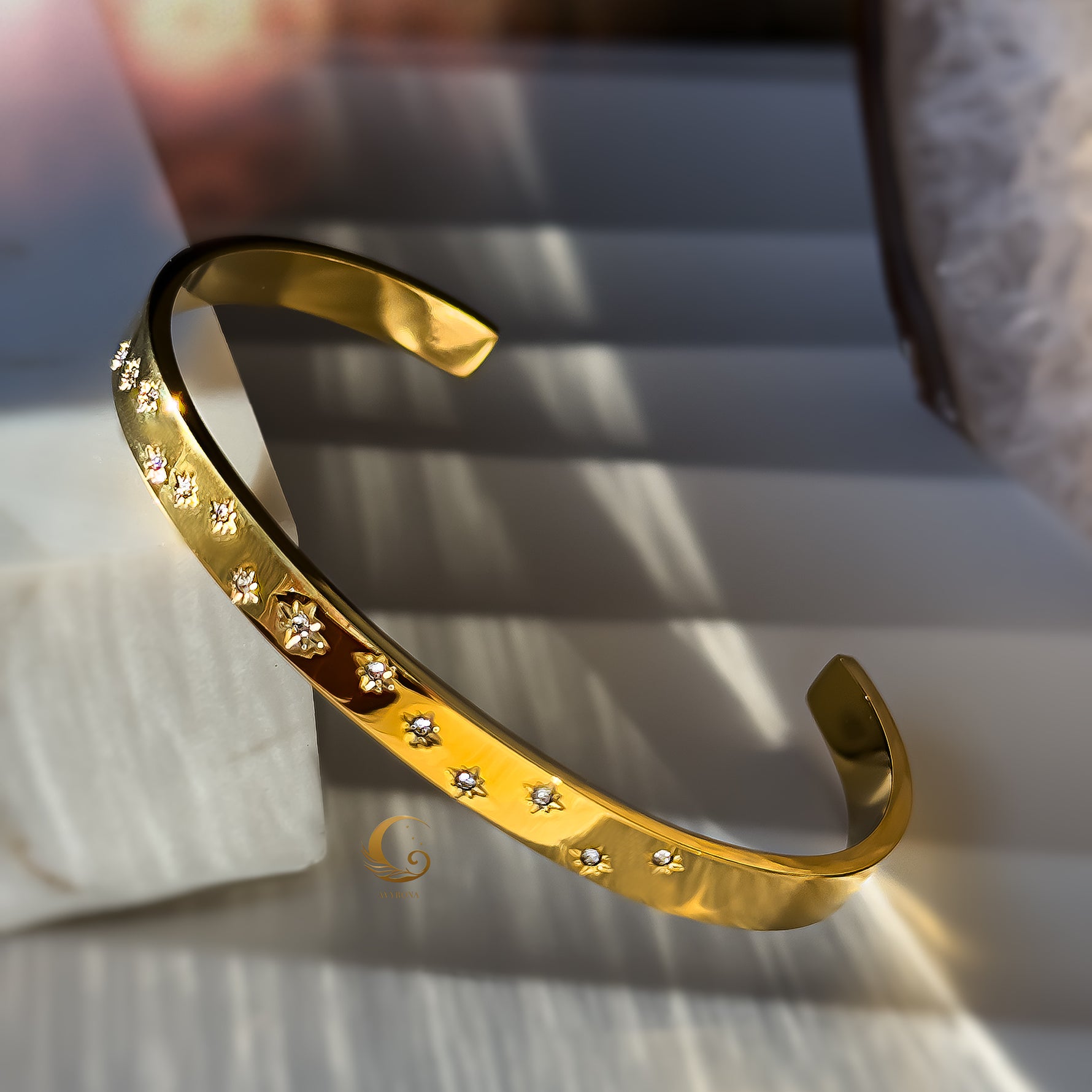 Single golden cuff bracelet with delicate engraved stars and stones, styled on a soft white stone.