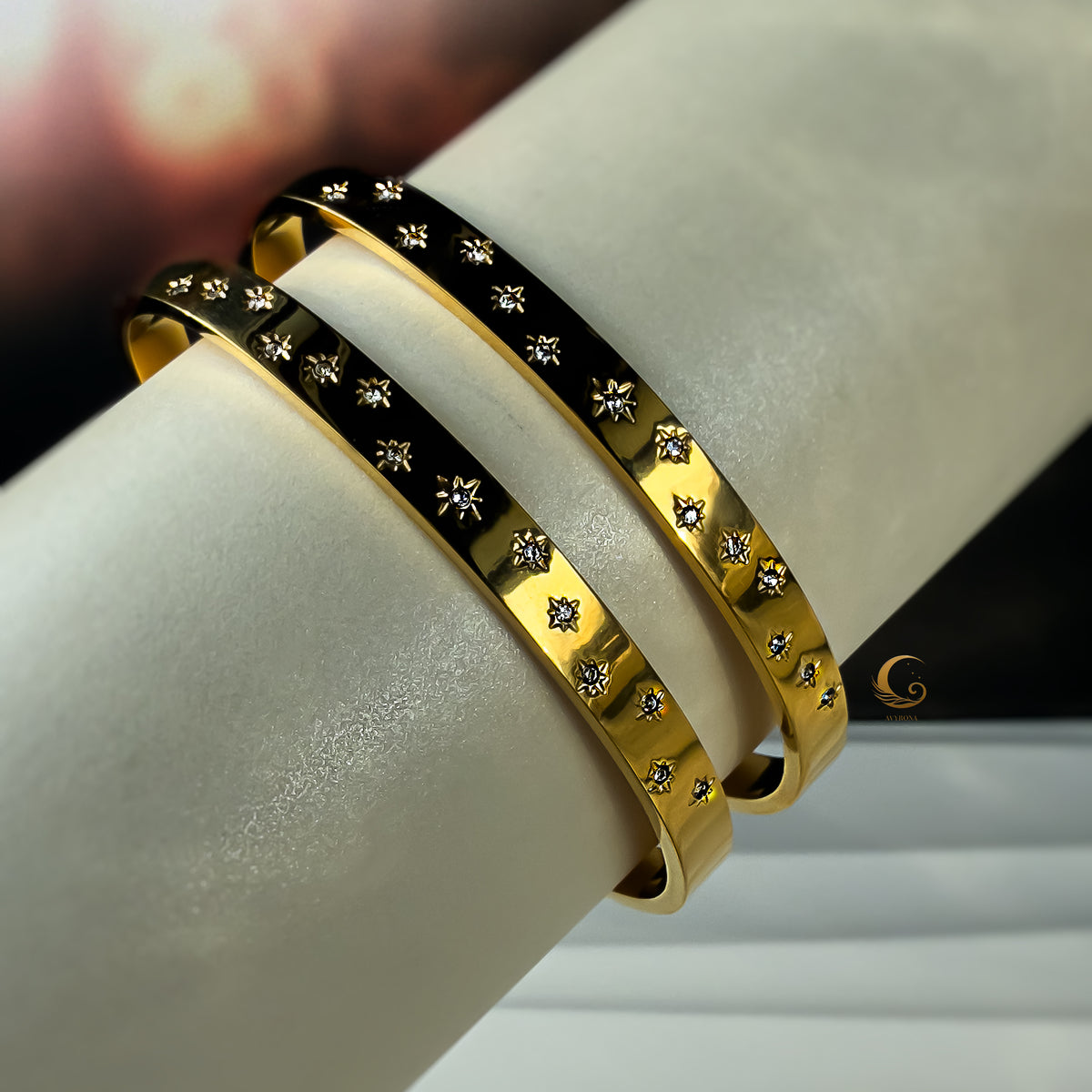Close-up of two gold stainless steel bracelets with minimal star patterns, displayed on a light roll cushion.