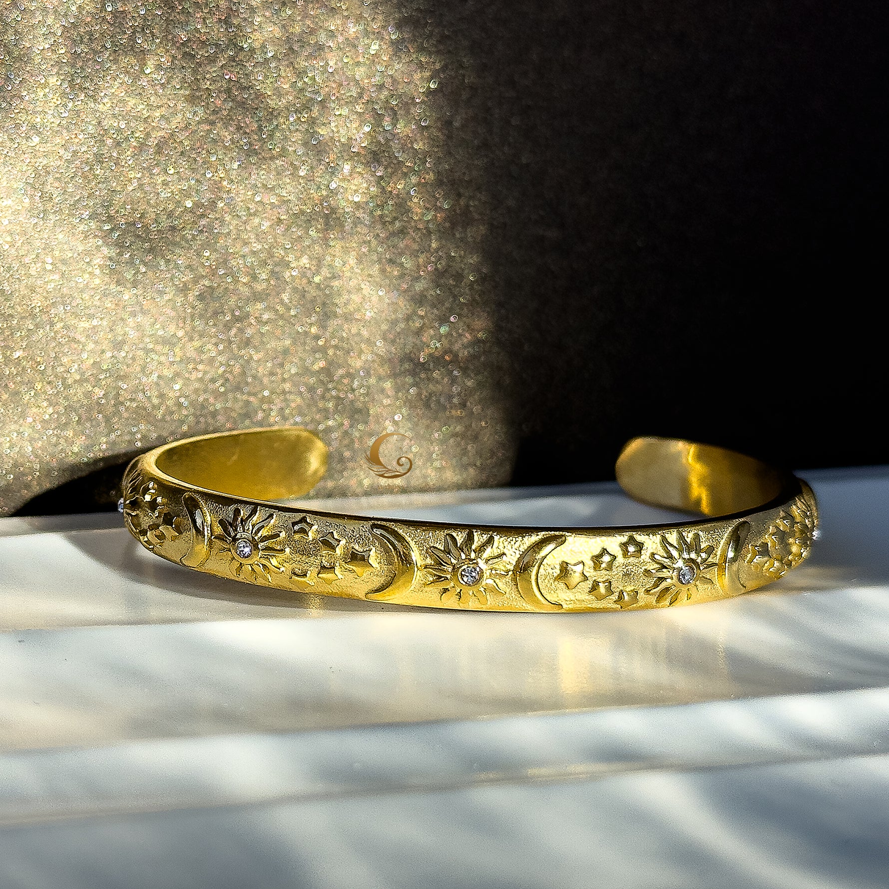 Gold cuff bracelet engraved with sun, moon, and star motifs, set with tiny cubic zirconia stones. Photographed against a soft glitter background with celestial shadows.