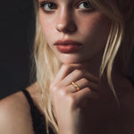 Close-up of a woman with blonde hair and a gold ring on her finger, against a dark background.