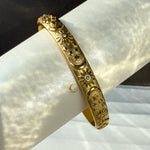  Side view of a gold-plated open bangle bracelet featuring engraved cosmic symbols—sun, crescent moons, and stars—on a soft rolled background with shimmering light.