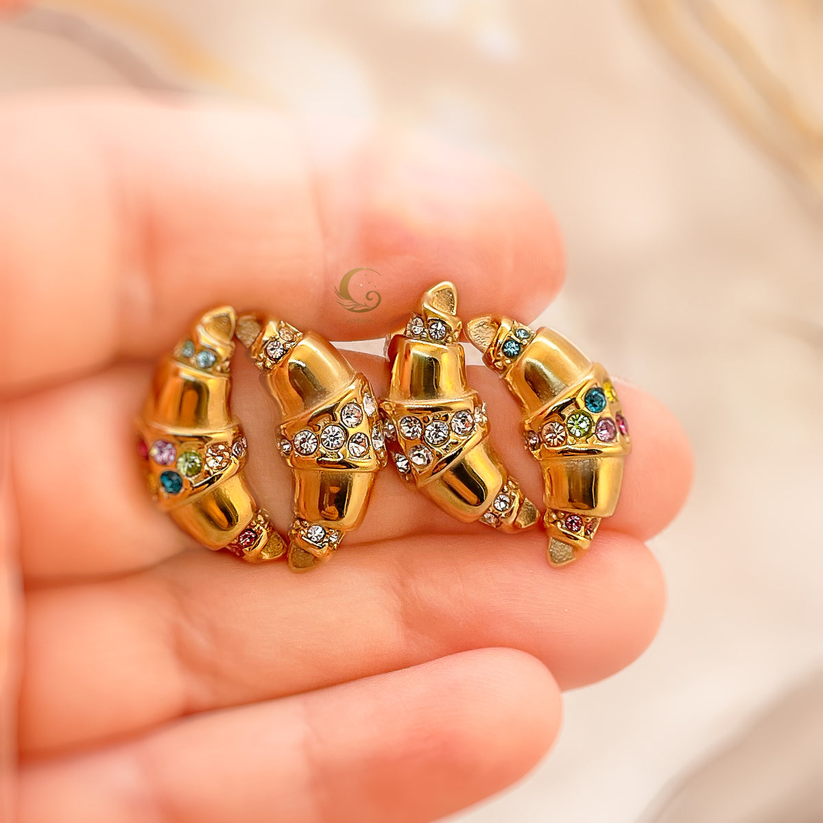  Hand holding four gold croissant-shaped earrings in clear and multicolored versions