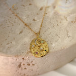 Gold-plated celestial pendant necklace with multicolor crystals, styled on creamy textured background