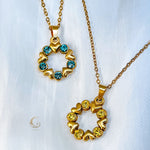 Two versions of the Elina pendant shown side by side – one with blue crystals, one with yellow crystals. Both are heart-shaped wreaths on gold chains.