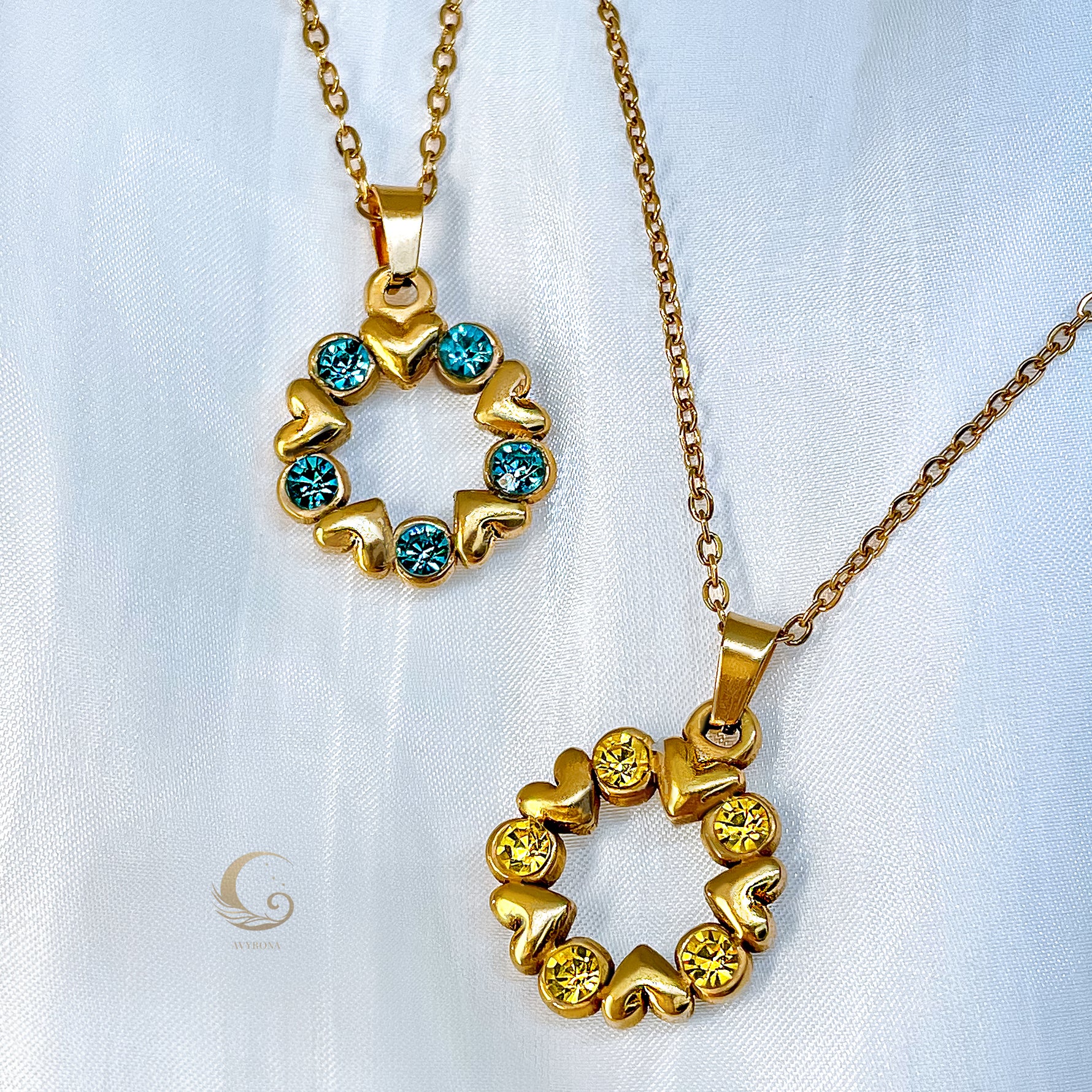 Two versions of the Elina pendant shown side by side – one with blue crystals, one with yellow crystals. Both are heart-shaped wreaths on gold chains.