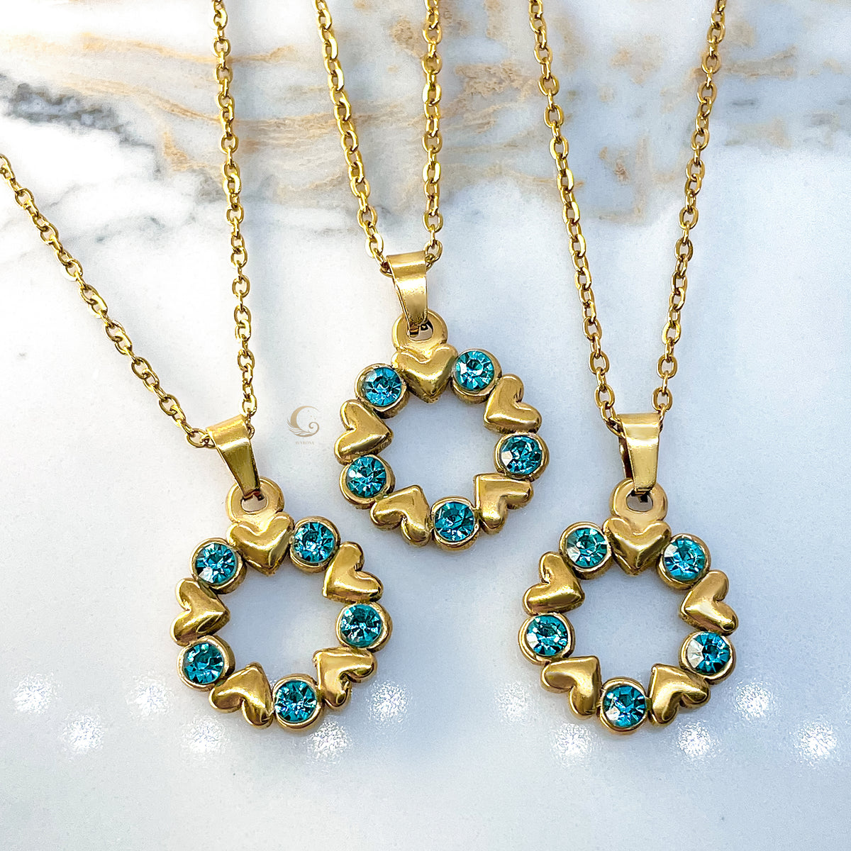 Three Elina necklaces displayed on a marble background. Each pendant shows heart-shaped gold accents paired with bright blue crystals forming a wreath.