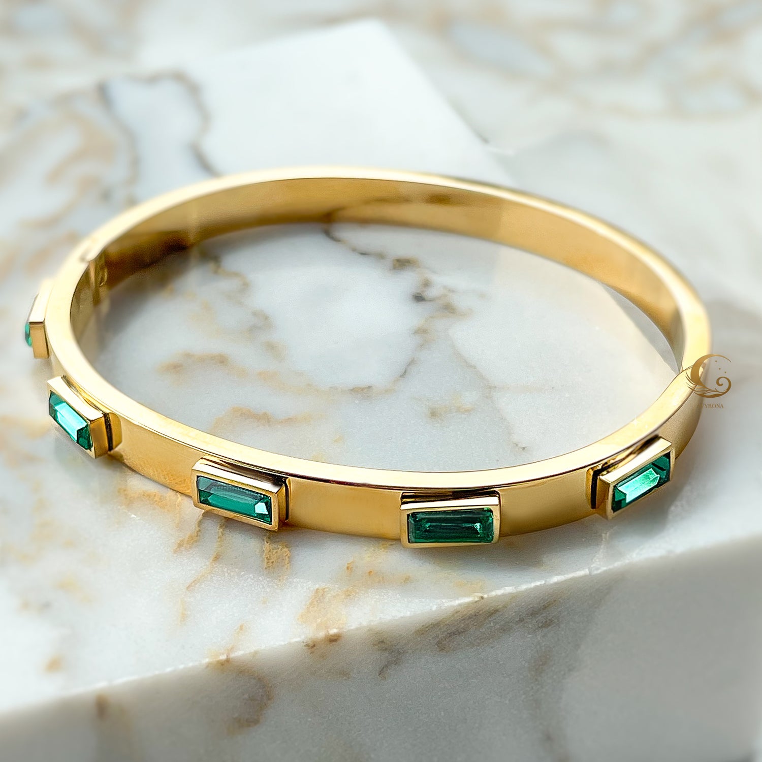 Top view of gold-plated stainless steel bangle bracelet with green crystals, photographed on a marble background.