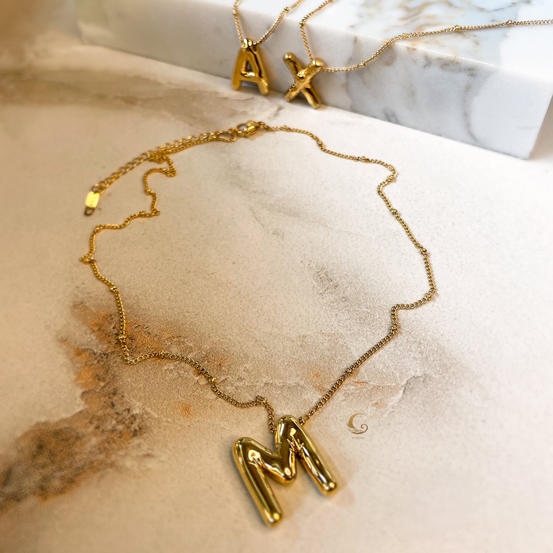 Gold balloon letter necklaces featuring the letters “A”, “X”, and “M”, displayed on a soft marble background.