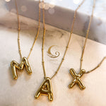 Close-up of gold balloon letter necklaces spelling “MAX”, vertically displayed on soft background with Avirona logo.