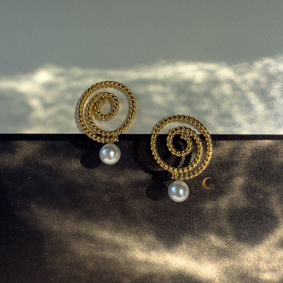 Gold Thaleia earrings with pearl-like drops on black and gold surface, glowing in contrast