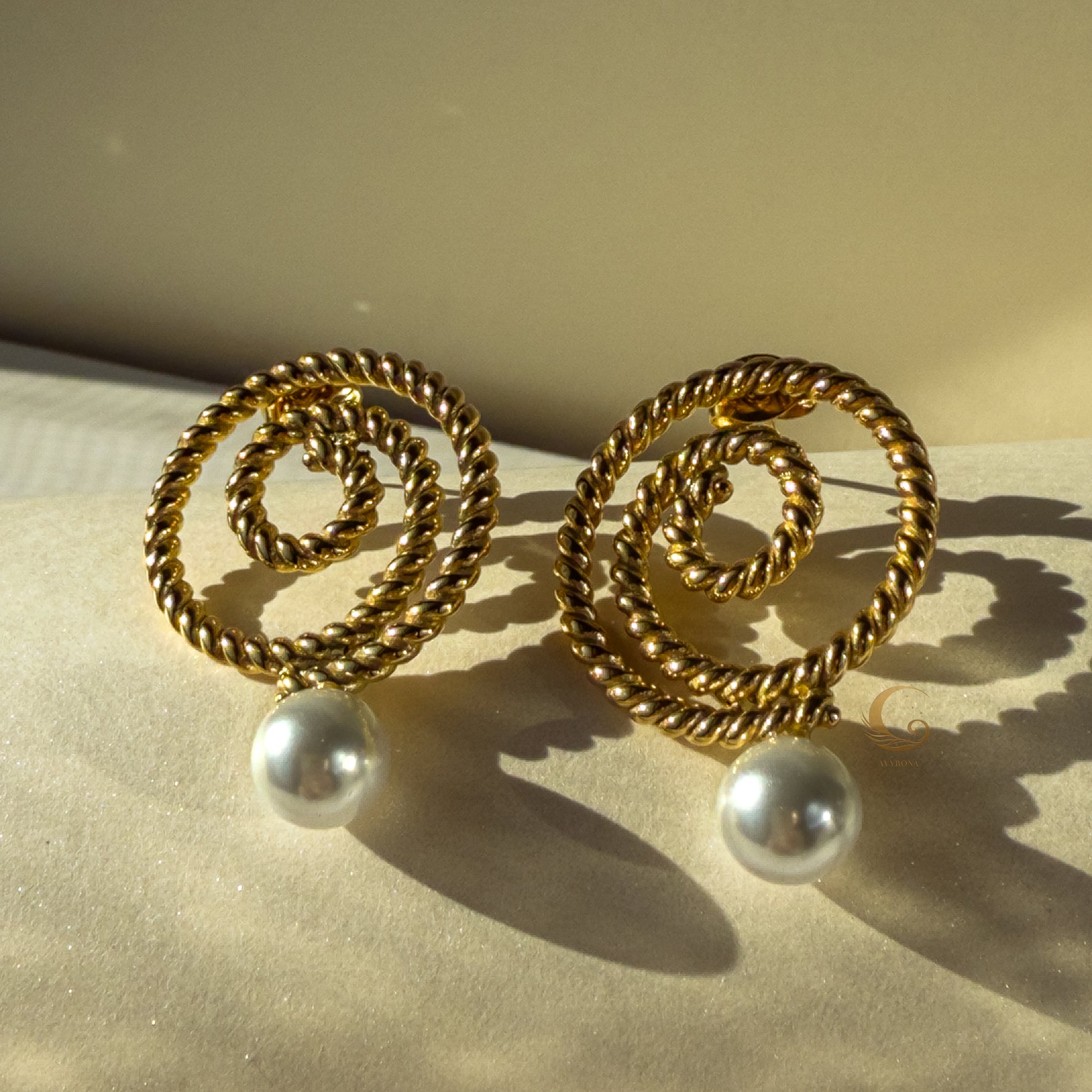 Thaleia gold spiral earrings casting elegant shadows, finished with pearl-like ends