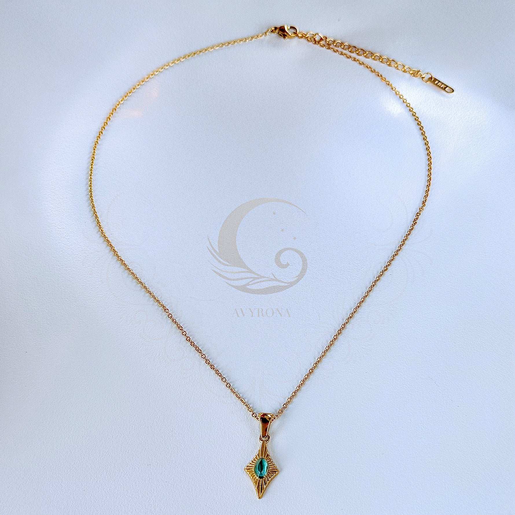 Full view of Guardian Eye necklace with blue cat eye stone pendant on a gold chain with extender, laid flat on Avyrona branded background.