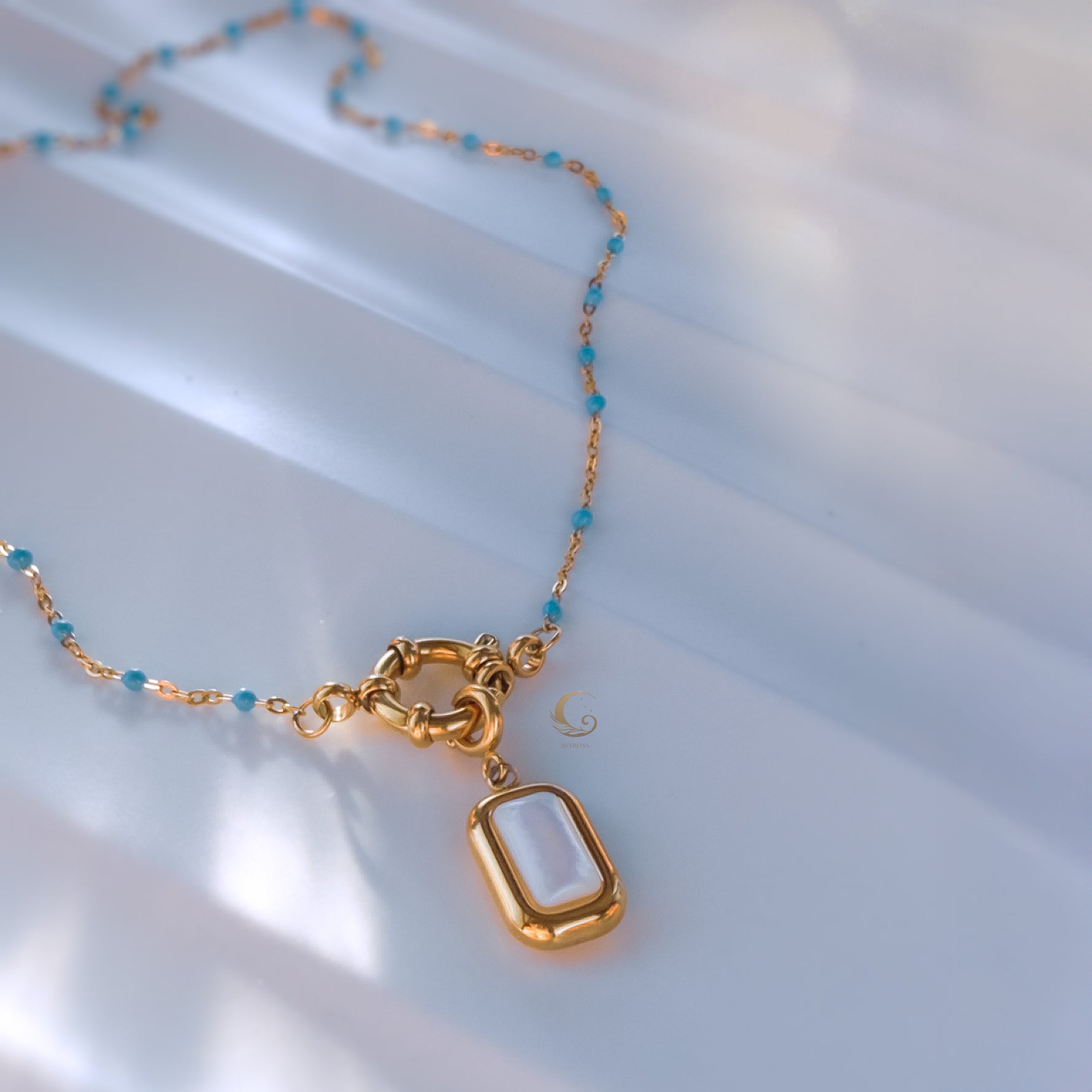 Sky blue version of the Liora necklace laid out flat with golden toggle clasp and rectangular mother of pearl pendant reflecting soft light.