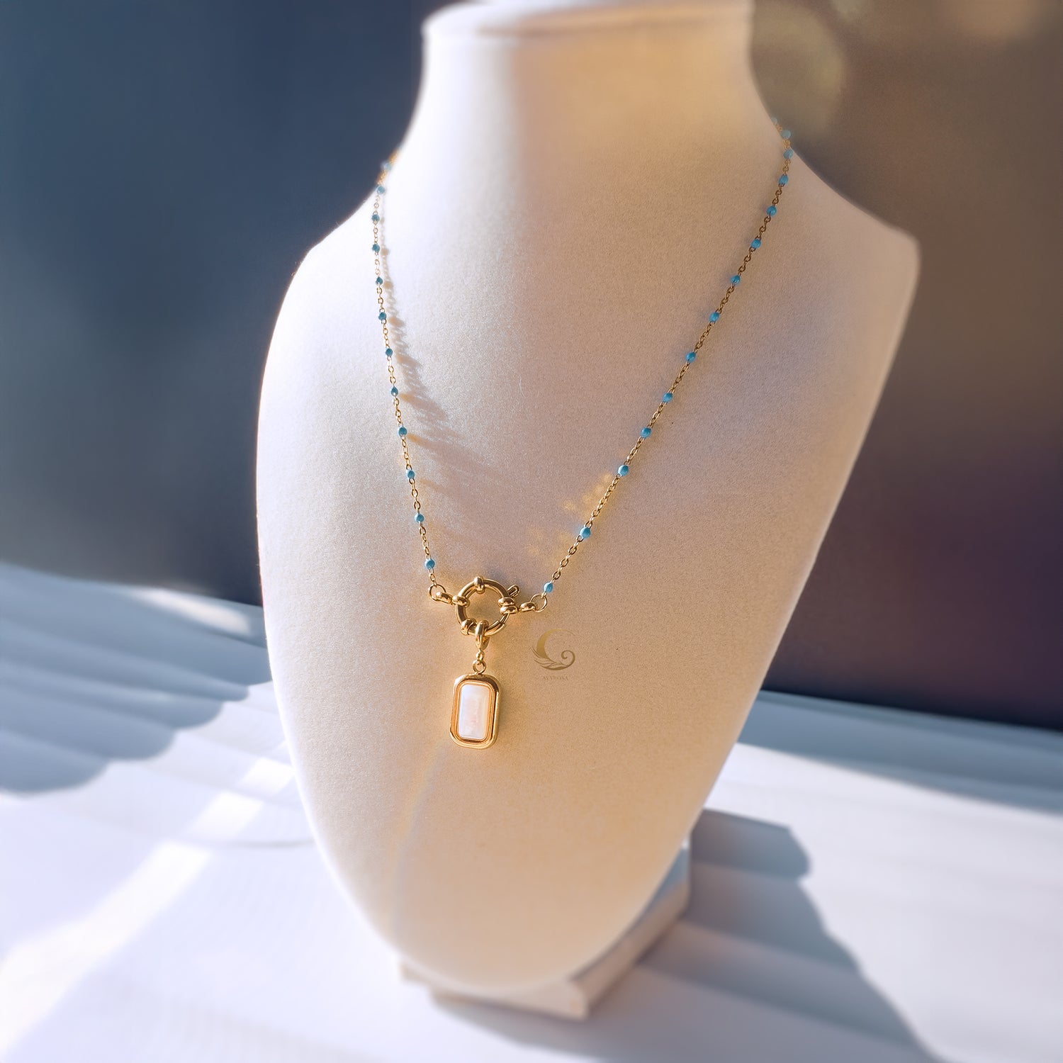  Liora beaded necklace displayed on a mannequin bust, showcasing the light blue beads, gold toggle clasp, and luminous mother of pearl pendant.