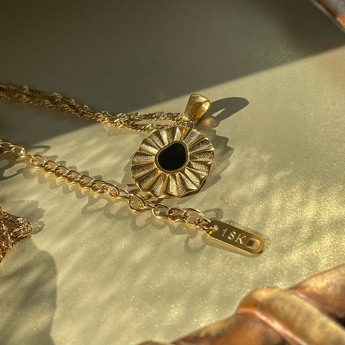 Liore Black Flower Necklace photographed on a shiny gold background, showing the floral pendant, the beginning of the chain, and the chain extension with a small gold 18K tag.