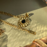 Liore Black Flower Necklace photographed on a shiny gold background, showing the floral pendant, the beginning of the chain, and the chain extension with a small gold 18K tag.