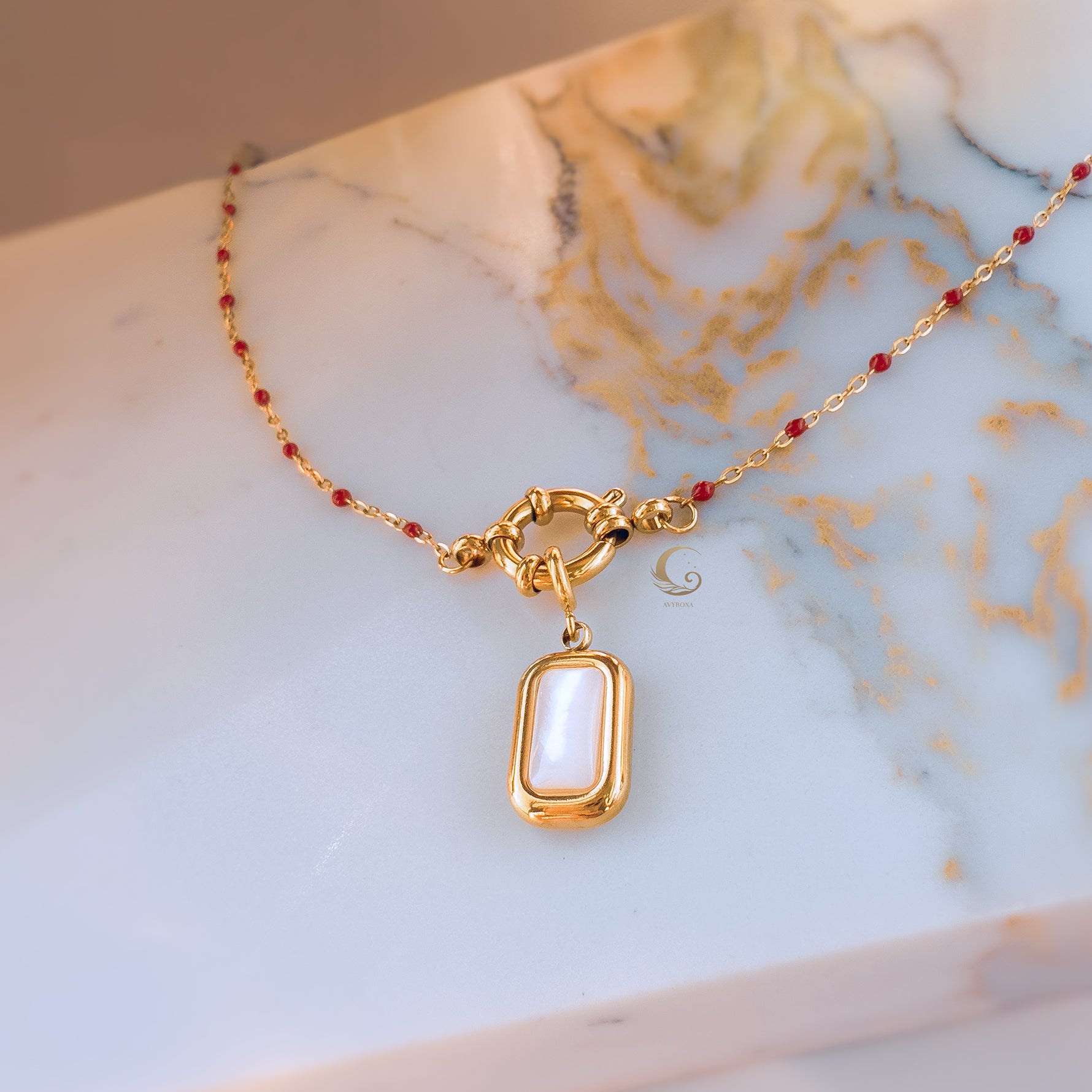 Red and gold necklace with toggle clasp and rectangular mother of pearl pendant on soft marble backdrop, angled lighting