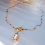 Full view of red beaded necklace with gold toggle clasp and mother of pearl pendant arranged in oval shape on white satin