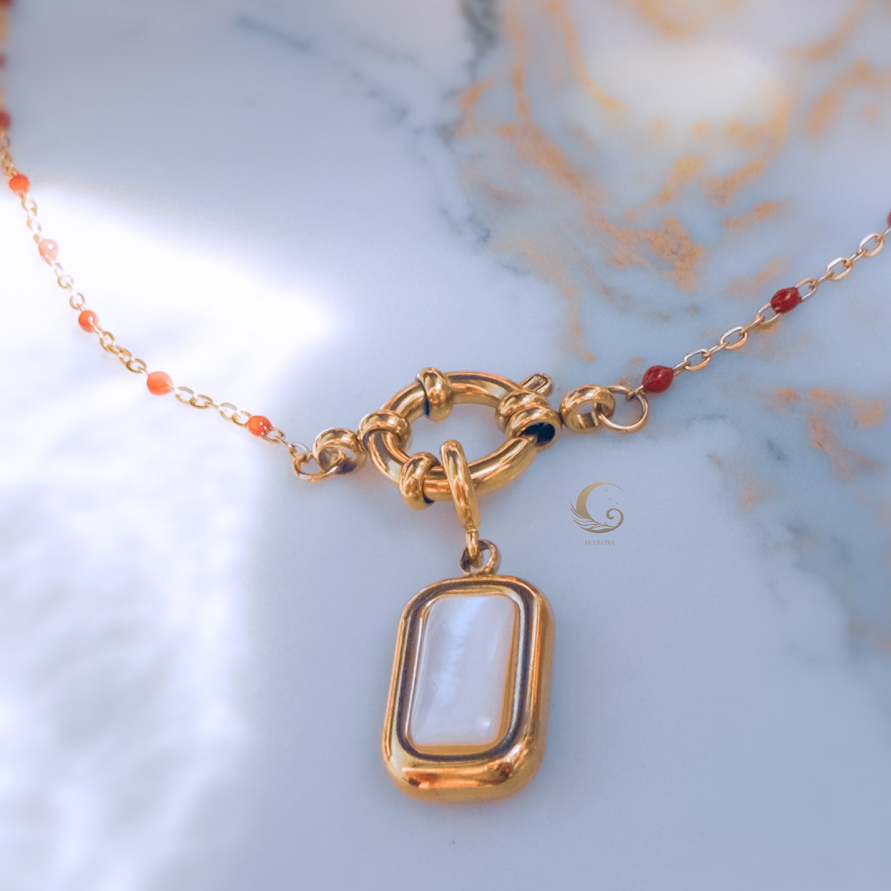  Close-up of gold toggle clasp with rectangular mother of pearl pendant and red beaded chain on marble background, soft light