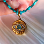Detailed shot of the gold Evil Eye pendant with turquoise and clear stones, on blue-green beaded chain.