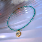 Full view of the Marisel turquoise Evil Eye necklace laid on soft white fabric with gold details and delicate beads.