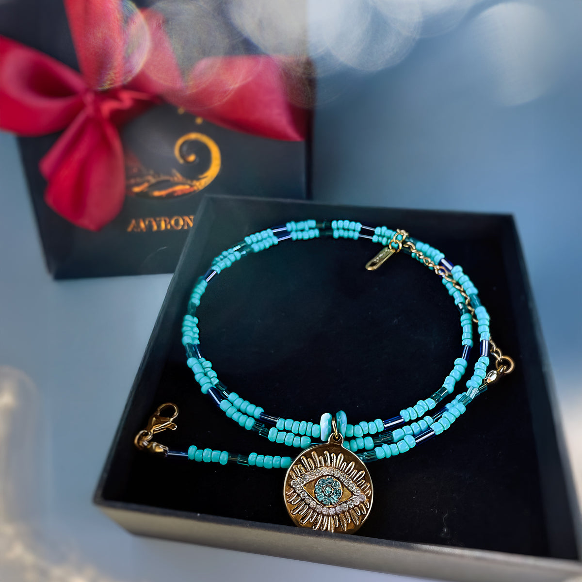 Turquoise beaded bracelet with gold evil eye charm on a black box with a red bow.