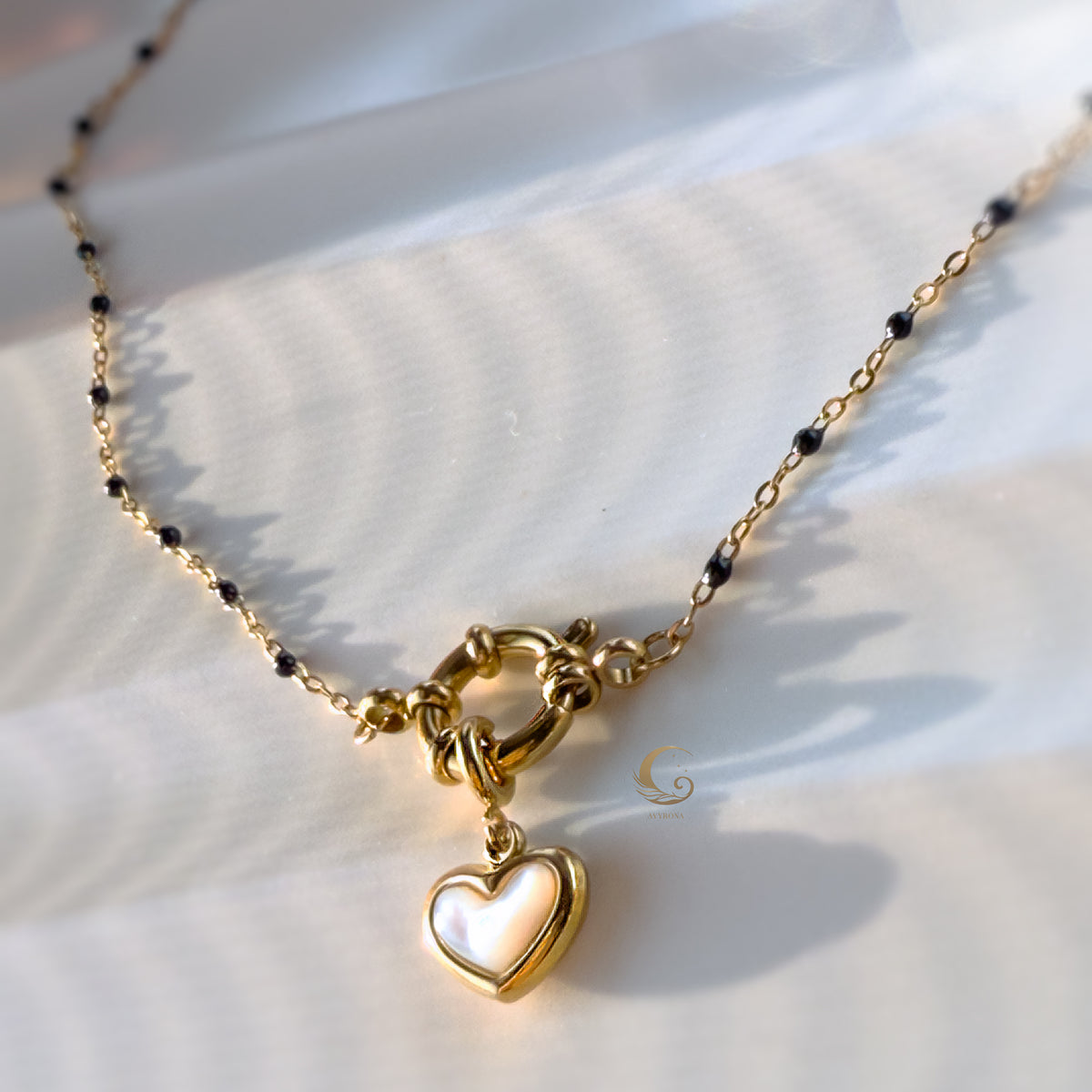 Close-up of gold heart pendant on navy beaded chain, lying on soft cream fabric with light reflections.