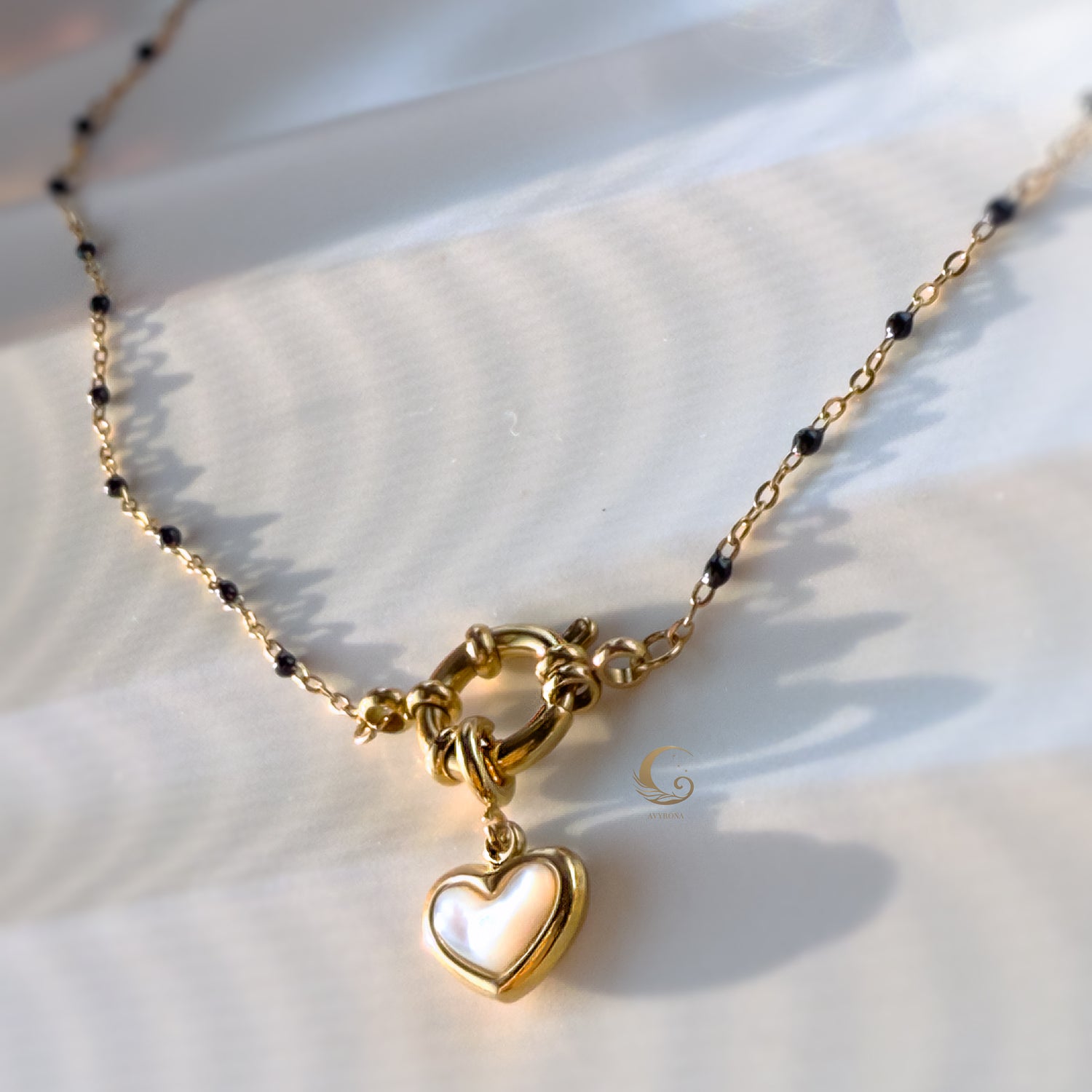 Close-up of gold heart pendant on navy beaded chain, lying on soft cream fabric with light reflections.