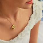 Model wearing a gold balloon-style “M” letter necklace on satellite chain, dressed in a white textured top.