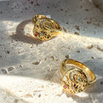 Artistic top view of two celestial gold rings with stars, moon, and evil eye symbols, placed on textured stone surface. Magic-inspired statement rings.