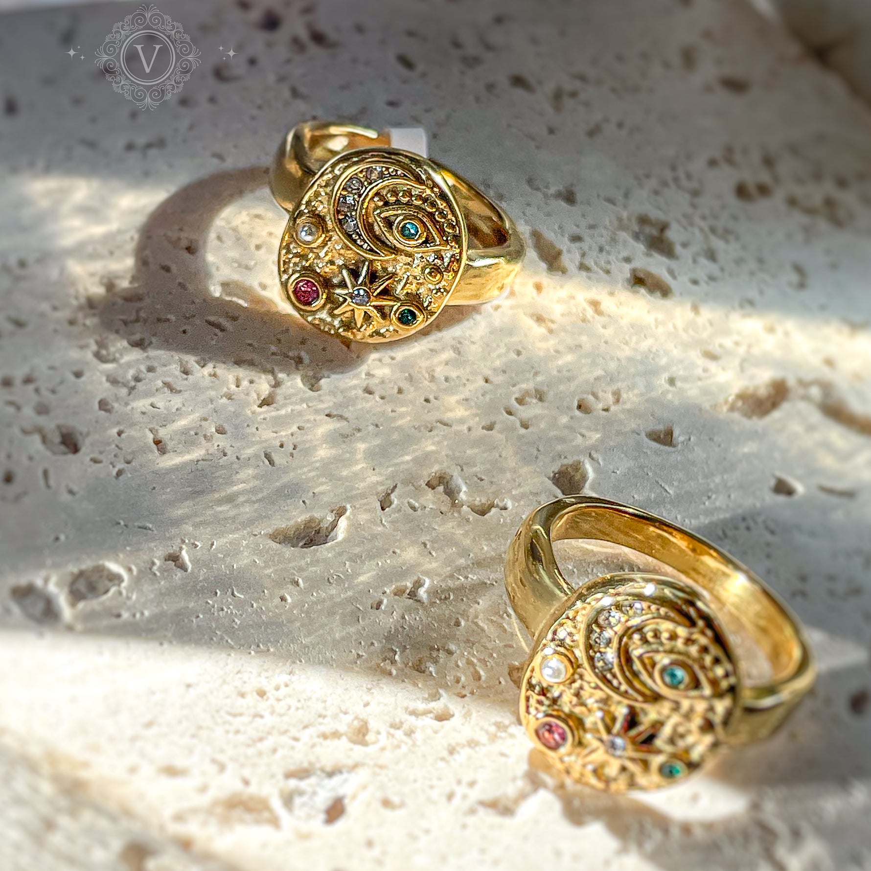 Artistic top view of two celestial gold rings with stars, moon, and evil eye symbols, placed on textured stone surface. Magic-inspired statement rings.