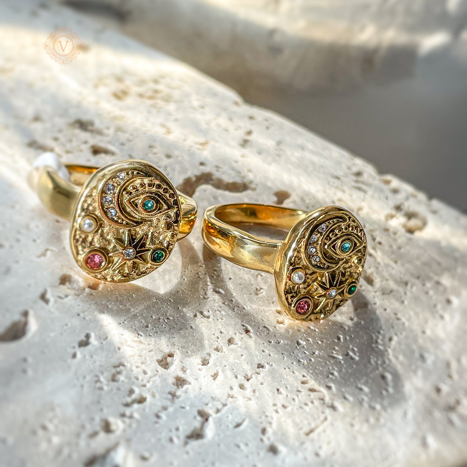 Two gold celestial rings with engraved moon and evil eye design, lying on beige stone. Detailed craftsmanship and textured surface shine in natural light.