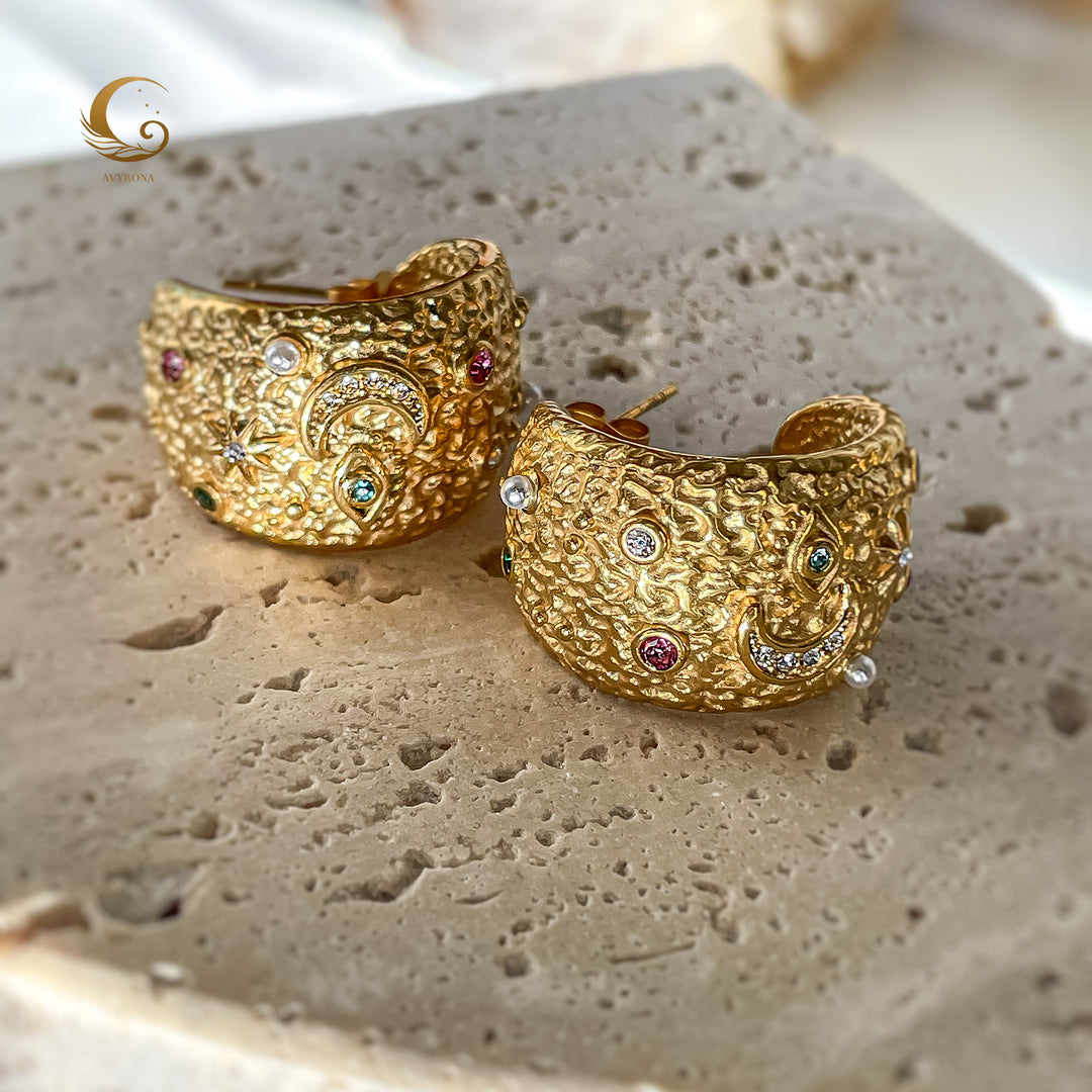 Close-up of large textured gold hoop earrings with moon, star, and evil eye symbols, placed on stone surface, soft natural light.