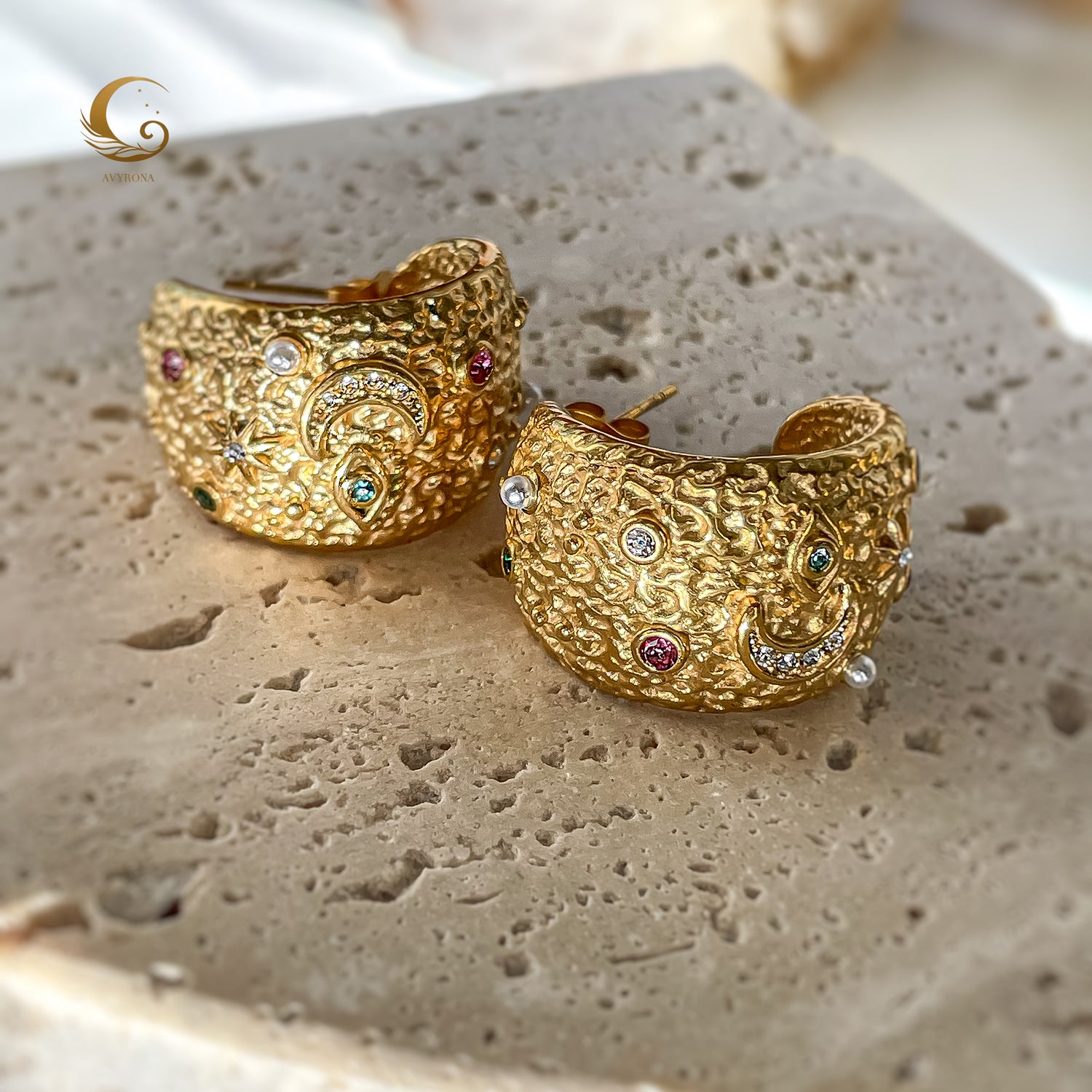 Close-up of large textured gold hoop earrings with moon, star, and evil eye symbols, placed on stone surface, soft natural light.