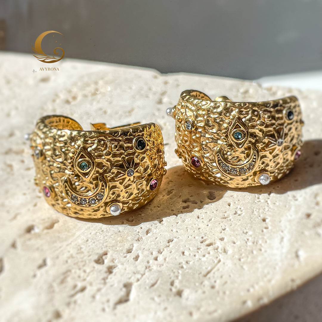  Pair of celestial gold hoop earrings displayed on beige stone, featuring detailed star, crescent moon, and eye motifs with colorful crystals.