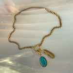 Gold cable chain necklace with feather and turquoise charm laid flat on silky background