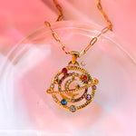 Gold celestial pendant with colorful stones and crescent moon on blush stone-like surface, AVYRONA jewelry.