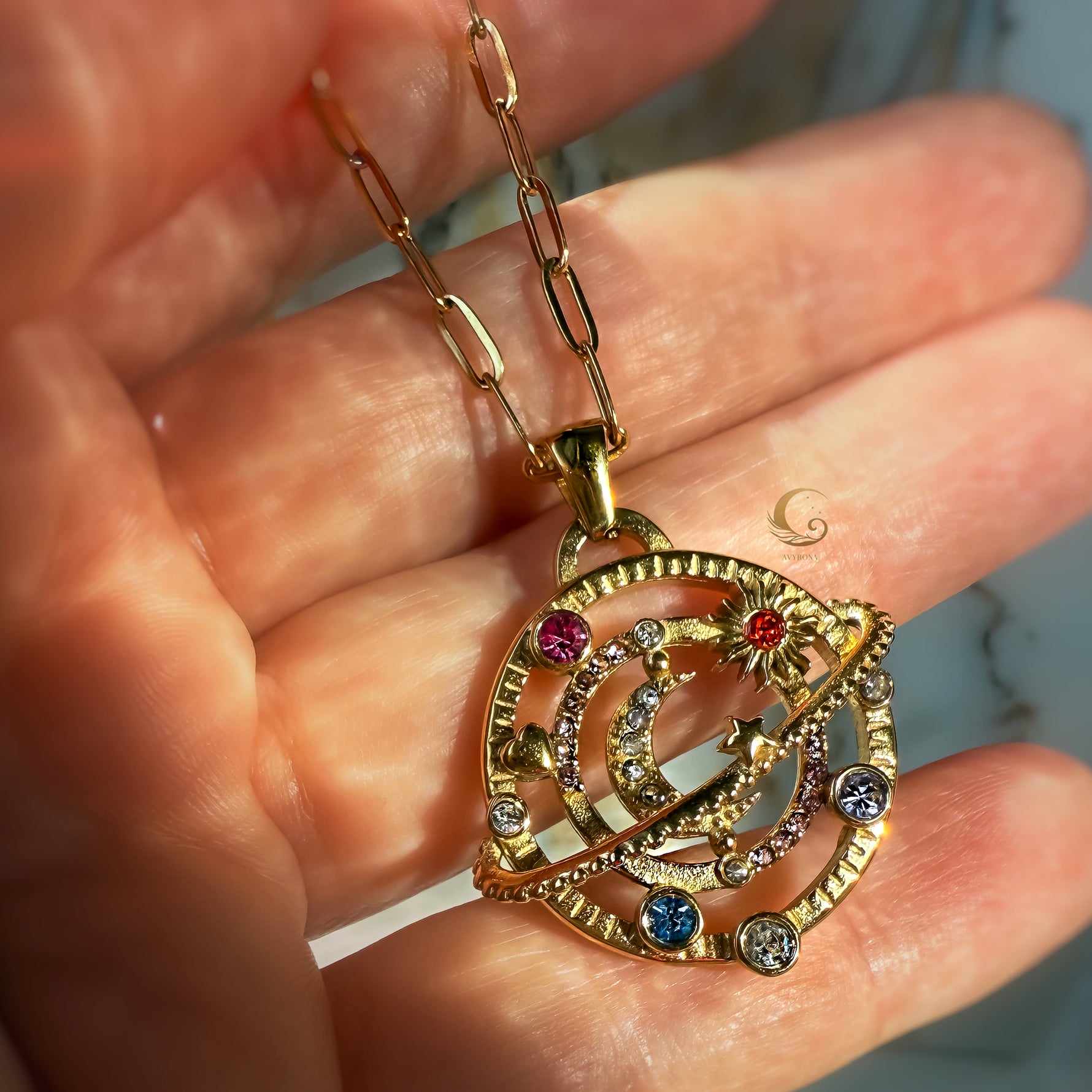 Close-up of AVYRONA gold moon and planet pendant with colorful rhinestones, held by hand on stone background.