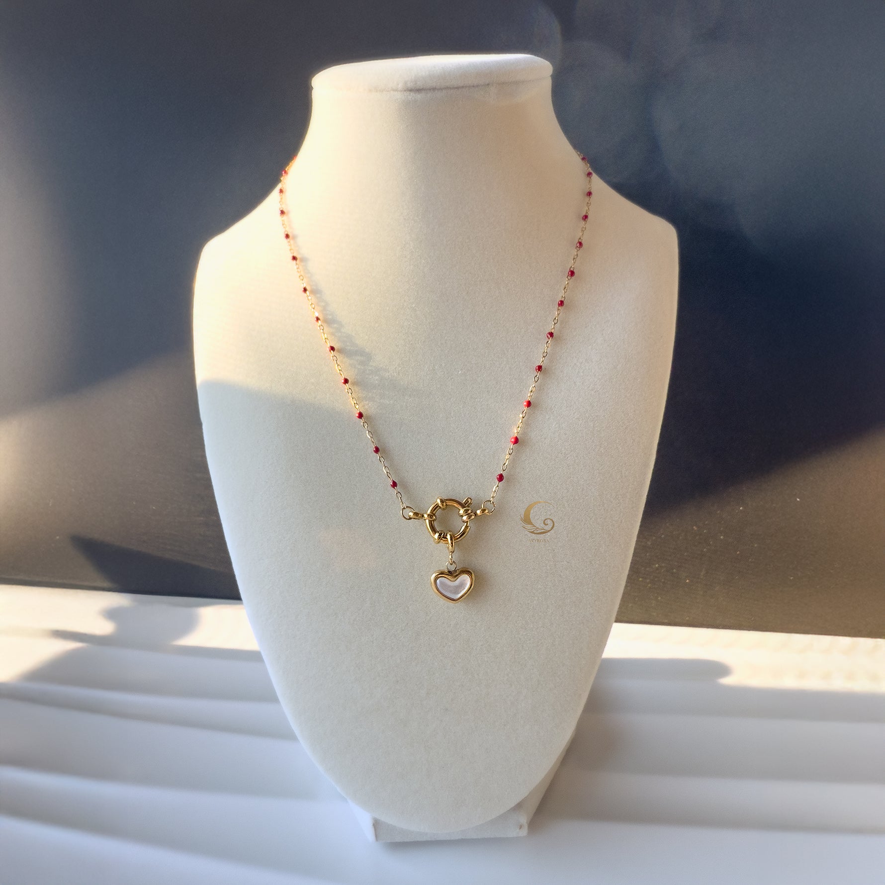 Rhea necklace with red beads and heart charm shown on white display bust in natural light