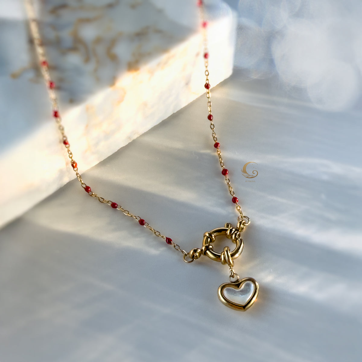 Close-up of red beaded necklace with toggle clasp and gold-framed heart pendant lying on pearlescent background