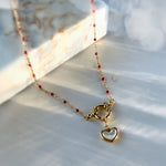 Close-up of red beaded necklace with toggle clasp and gold-framed heart pendant lying on pearlescent background