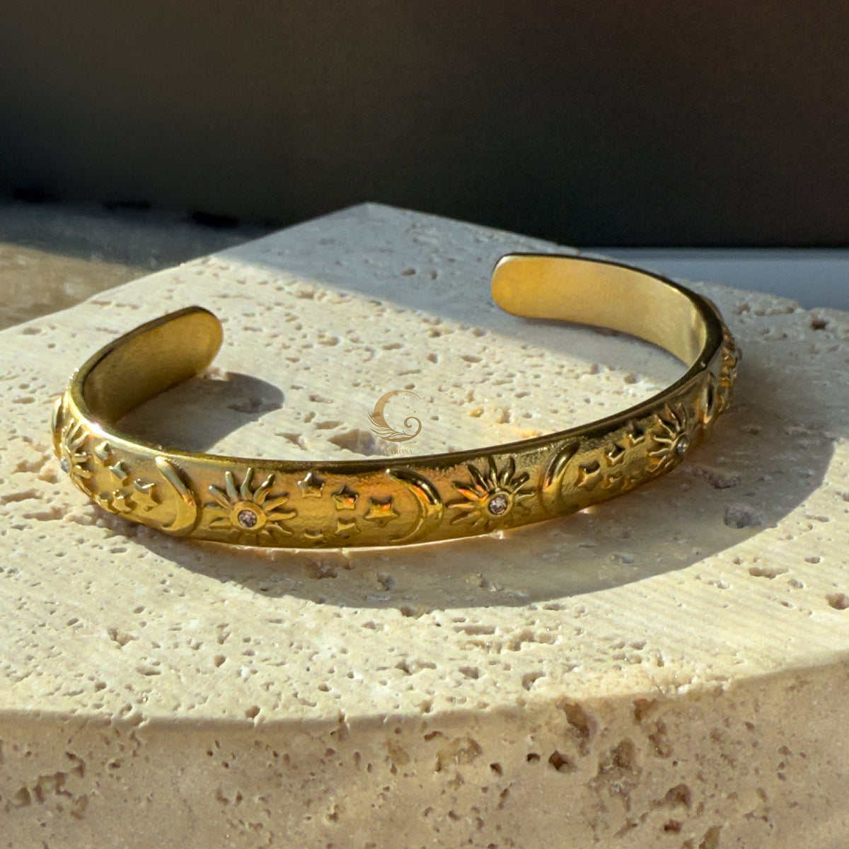 Side view of open gold cuff bracelet with celestial engravings, set against textured stone for an earthy, elegant contrast.