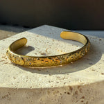 Side view of open gold cuff bracelet with celestial engravings, set against textured stone for an earthy, elegant contrast.