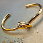 Gold open cuff bracelet with symbolic knot detail, photographed on shimmering surface – Avyrona Earth Collection